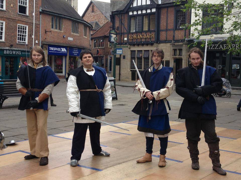 Four men, in matching tabards, padded coats and with swords, arrayed on a stage in the middle of a town square.