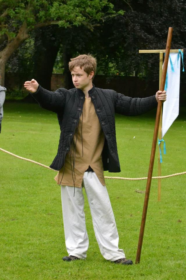 Man in padded coat, propped against a quarterstaff, officiating an unseen bout